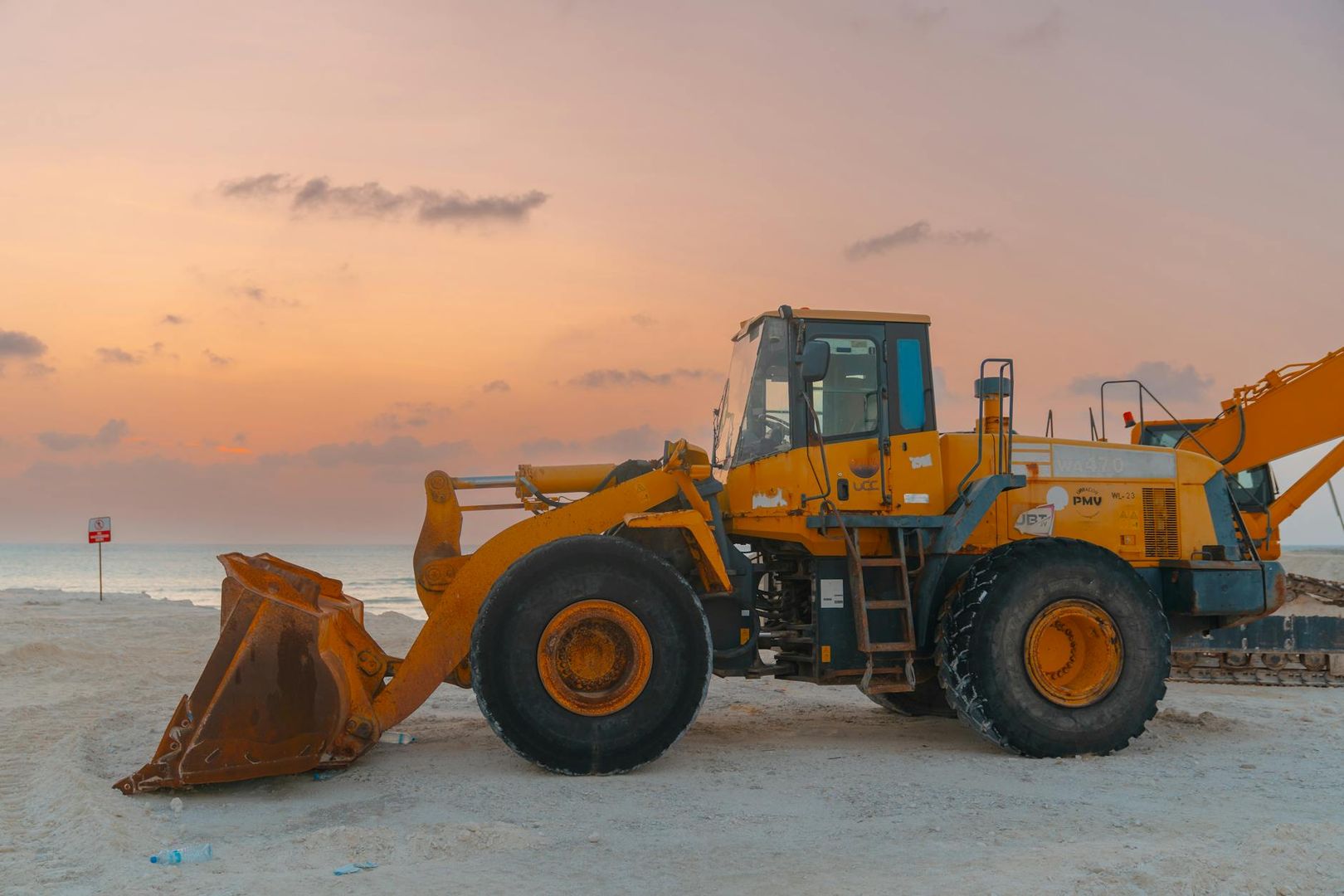 Gelber Radlader auf Sandboden vor einem farbenprächtigen Abendhimmel am Meer.